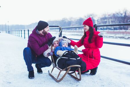 Beautiful young couple near their kids, sitting on sleigh. Lovely girl and cute boy muffled in warm cover on wooden sleigh with their parents sitting near. Snowy city view on backgroundの写真素材