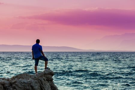 Yung man standing on a rock and looking at the sea sunsetの写真素材