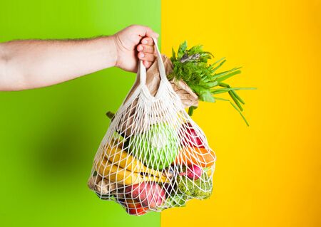 Zero waste shopping. Man hand holds cotton bag full of vegetables on green and yellow background. Produce in paper bag. Place for text, vegan foodの写真素材