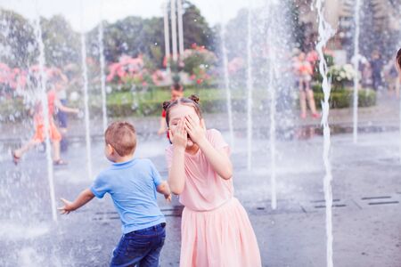 Little girl in a wet pink dress is standing in the middle of the fountain and is covering her face with her handsの写真素材