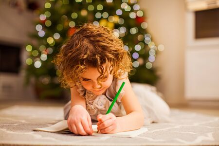 Cute little girl with curly hair in beautiful dress , laying on a floor, writing in her notebook with green pencil. Christmas tree with lights on a background, bluredの写真素材