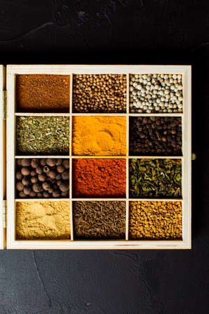 Wooden storage for spices. Box divided into cells, filled with diverse dry herbs and spices. Colorful and diverse condiments, top view on black backgroundの写真素材