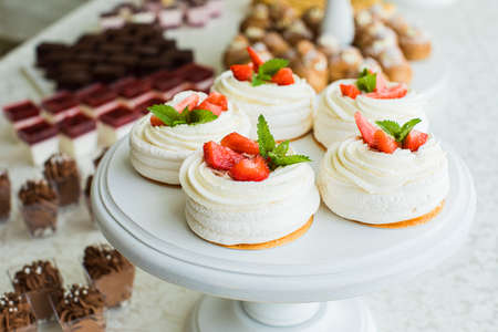 Pavlova cakes with delicate white cream and strawberries are laid out on a high white stand and many other sweets in the backgroundの写真素材