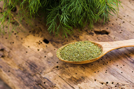 From above view fresh dill sprigs with wooden spoon full of dried and milled dill on wooden board. Horizontal closeup photo of homemade dill seasoning on old crannied surfaceの写真素材