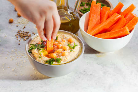 Closeup womans hand dipping piece of carrot into bowl with hummus paste. Bowl with carrots and glass jar of olive oil placed near. Party snack. White background with spilled sezame seeds, bluredの写真素材
