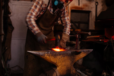 The blacksmith manually forging the molten metal on the anvil in smithy with spark fireworks.