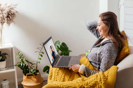 Cheerful girl sitting in armchair chatting with friends onlineの写真素材