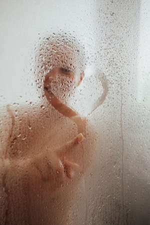 Cheerful woman drawing heart on condensed glass doorの写真素材