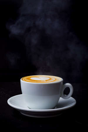 Cappuccino with steam and heart love latte art in white cup on the black tableの写真素材