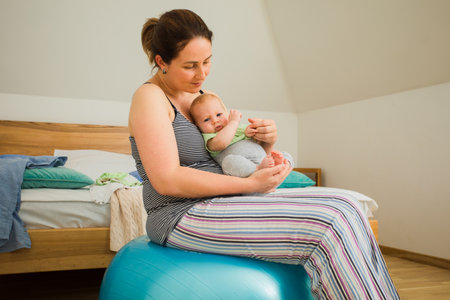 Mother holding and rocking baby sitting on fitballの写真素材
