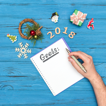 notebook with GOALS text and mans hand holding pen above with numbers 2018 and new year ornaments
