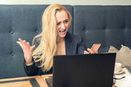 Angry dissatisfied young woman looking on the smartphone on phone in front of the laptop, having...
