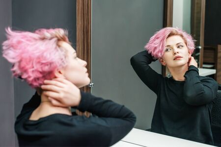 Beautiful hairstyle of young woman looking in the mirror after dying hair and making highlights in beauty salon. Close up short pink hairdoの写真素材