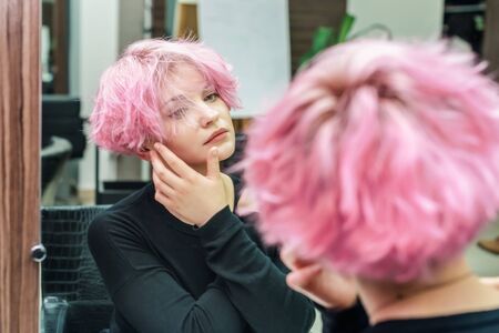 Beautiful hairstyle of young woman looking in the mirror after dying hair and making highlights in beauty salon. Close up short pink hairdoの写真素材
