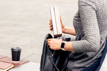 Female student puts books into into black backpack close up. Hands of woman student is putting books into backpack outdoors. Student life concept.の写真素材