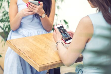 Two young women are looking at smart phone at cafe outdoors. Women sits in the street cafe with smartphone. Girl looks at herself on the smartphone.の写真素材