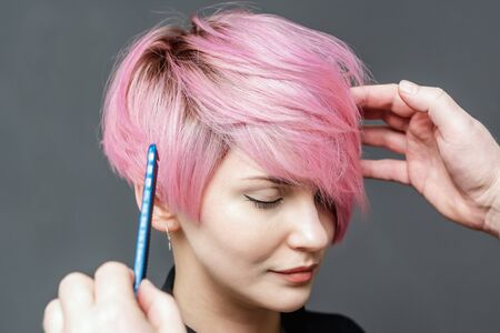 Hands of hairdresser checks short pink hair of girl on gray background close up. Hairdresser checks with comb short pink hairstyle of young woman.の写真素材