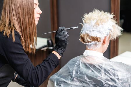 back view of hairdresser in black gloves are applying white color to woman hair. Hair coloring in white color at beauty salon.の写真素材