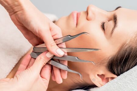 Cosmetologist holds tweezers for eyelash extensions near face of young woman, close up.
