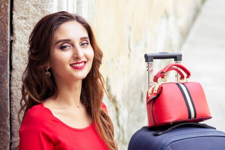 Portrait of beautiful smiling woman with suitcase in red dress over old wall.の写真素材