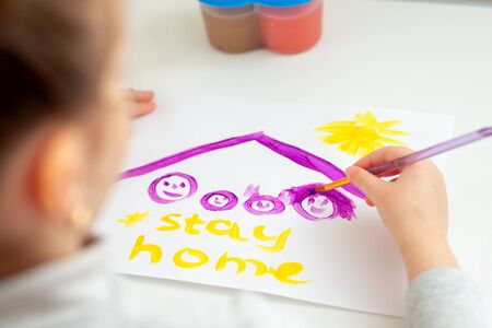 Child is drawing family under a roof with words Stay Home on sheet of paper.の写真素材