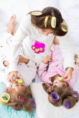 Mother and two daughters are putting cucumbers on eyes with curlers in hair lying on the bed in the bedroom. Stay home and family concept.の写真素材