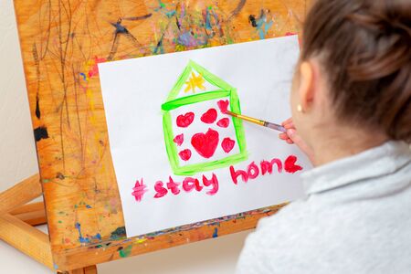 Stay Home concept. Child is drawing house with red hearts on the sheet of paper with a written...