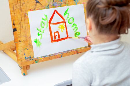 Child is drawing red house with words Stay Home on white sheet of paper at home. Stay Home concept.