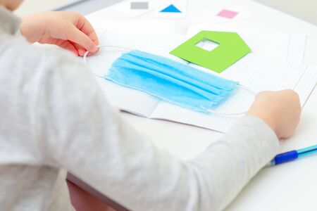 Children's hands holding a medical protective mask near paper green house over school notebook on the desk at home. Quarantine studying concept.の写真素材