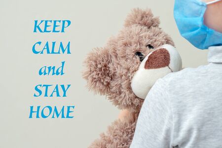 Child is holding toy bear in protective medical mask with words Keep Calm and Stay Home. Health care and virus protection concept.の写真素材