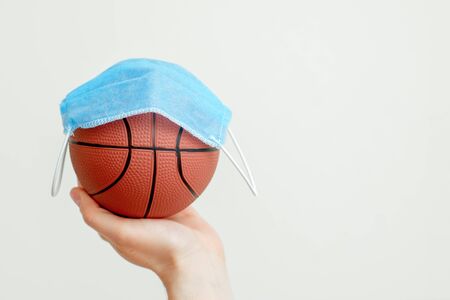 Basketball ball with medical mask on it in male hand on light background with copy space. Cancellation of sporting events.の写真素材