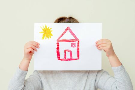 Closeup of Hands of child holding picture of red house with sun covering her face on yellow background. The child learns to draw.の写真素材