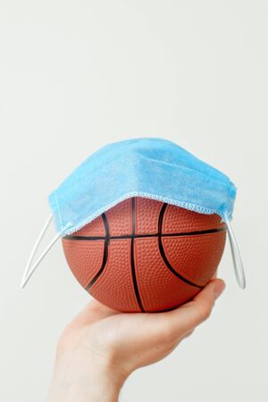 Closeup vertical image of protective medical mask over basketball ball on male hand on light background. Healthcare, sport concept.の写真素材