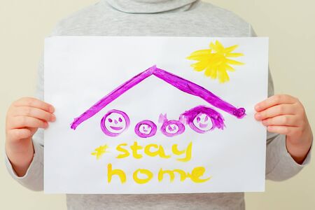 Hands of child holding a picture of family silhouette under the roof and words Stay Home on yellow background. Children's creativity concept.の写真素材