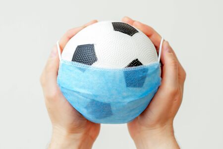 Closeup of soccer ball with medical protective mask in hands of man on light background during coronavirus. Coronavirus suspends sports world.の写真素材