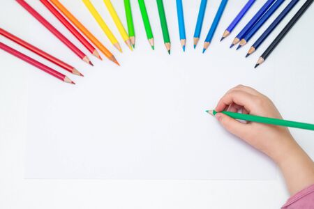 Top view of kid's left hand holding color pencil and drawing on white blank paper with the set of colour pencils. Copy space for text. Mockup.の写真素材