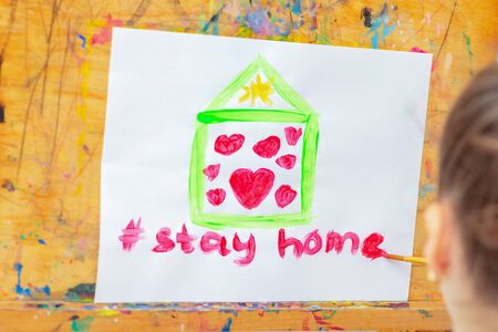 Child is drawing green house with red hearts and words Stay Home on sheet of paper during quarantine at home. Children's creativity.の写真素材