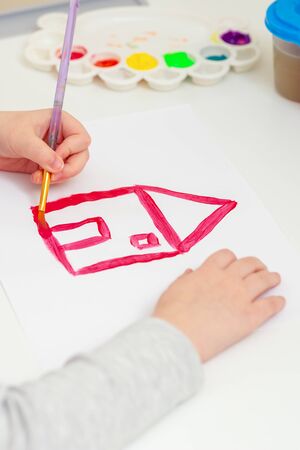 Side view vertical image of child hand with pencil is drawing picture of red house on white paper. Children's creativity.の写真素材