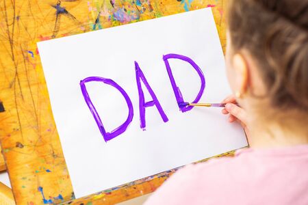 Hand of child drawing handwritten purple word Dad greeting card on white paper on easel. Family and Father's Day concept.の写真素材