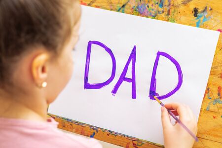 Child is drawing purple word Dad greeting card on white paper on easel. Father's Day concept.の写真素材