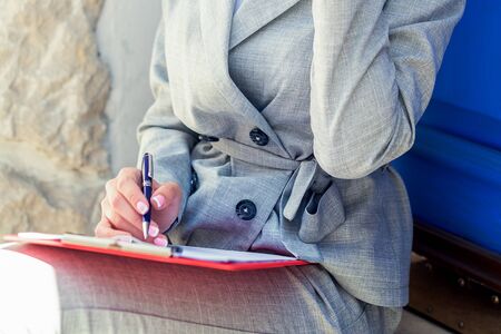 Close up of hand of woman writing on paper by pen on her knees outdoor.