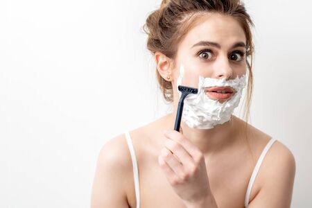 Beautiful young caucasian woman shaving her face by razor on white background. Pretty woman with shaving foam on her faceの写真素材