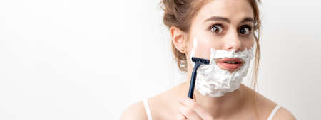 Beautiful young caucasian woman shaving her face by razor on white background. Pretty woman with shaving foam on her faceの写真素材