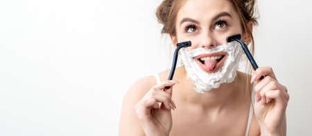 Beautiful young caucasian woman shaving her face by two razors with tongue out on white background.の写真素材