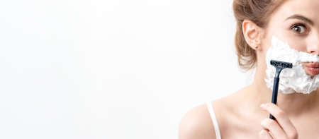Beautiful young caucasian woman shaving her face by razor on white background. Pretty woman with shaving foam on her faceの写真素材