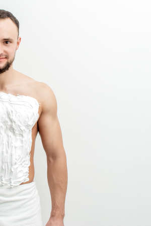 Young caucasian man with beard holds razor shaves his chest with white shaving foam on white...