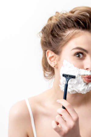 Beautiful young caucasian woman shaving her face by razor on white background. Pretty woman with shaving foam on her faceの写真素材