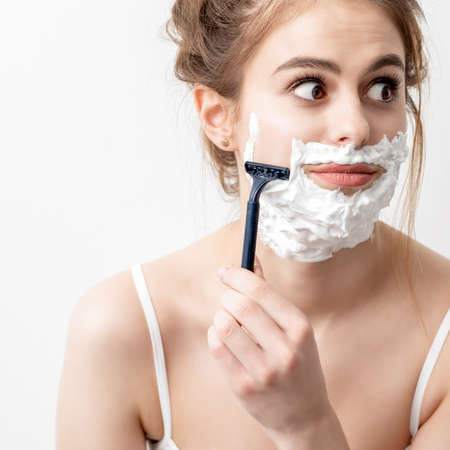 Beautiful young caucasian woman shaving her face by razor on white background. Pretty woman with shaving foam on her faceの写真素材