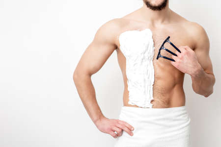 Young caucasian man with beard holds razor shaves his chest with white shaving foam on white background. Man shaving his torsoの写真素材