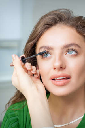 Makeup artist applies mascara on eyelash of young caucasian woman in beauty salon